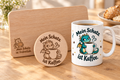 Drachen-Geschenkset mit weißer Motivtasse, graviertem Frühstücksbrettchen und rundem Holzuntersetzer auf einem Tisch.
